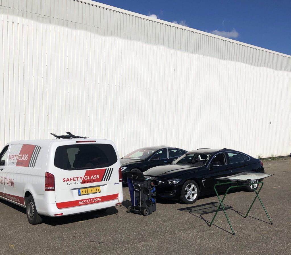 Servicebus van Safety Glass naast een auto waarvan de voorruit wordt vervangen, met een nieuwe ruit op een glasstandaard